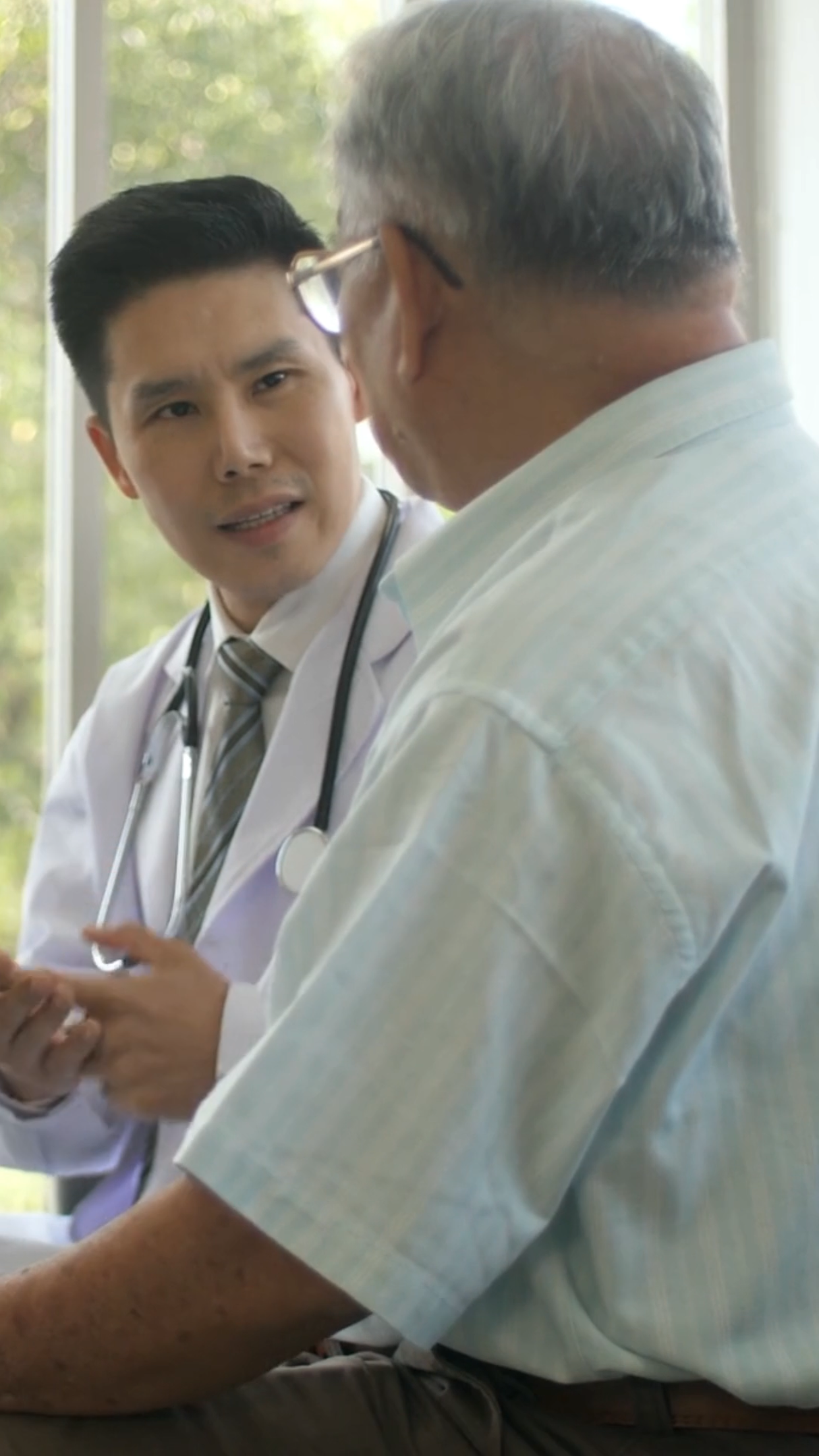 asian male primary care doctor sits down with older male patient in conversation